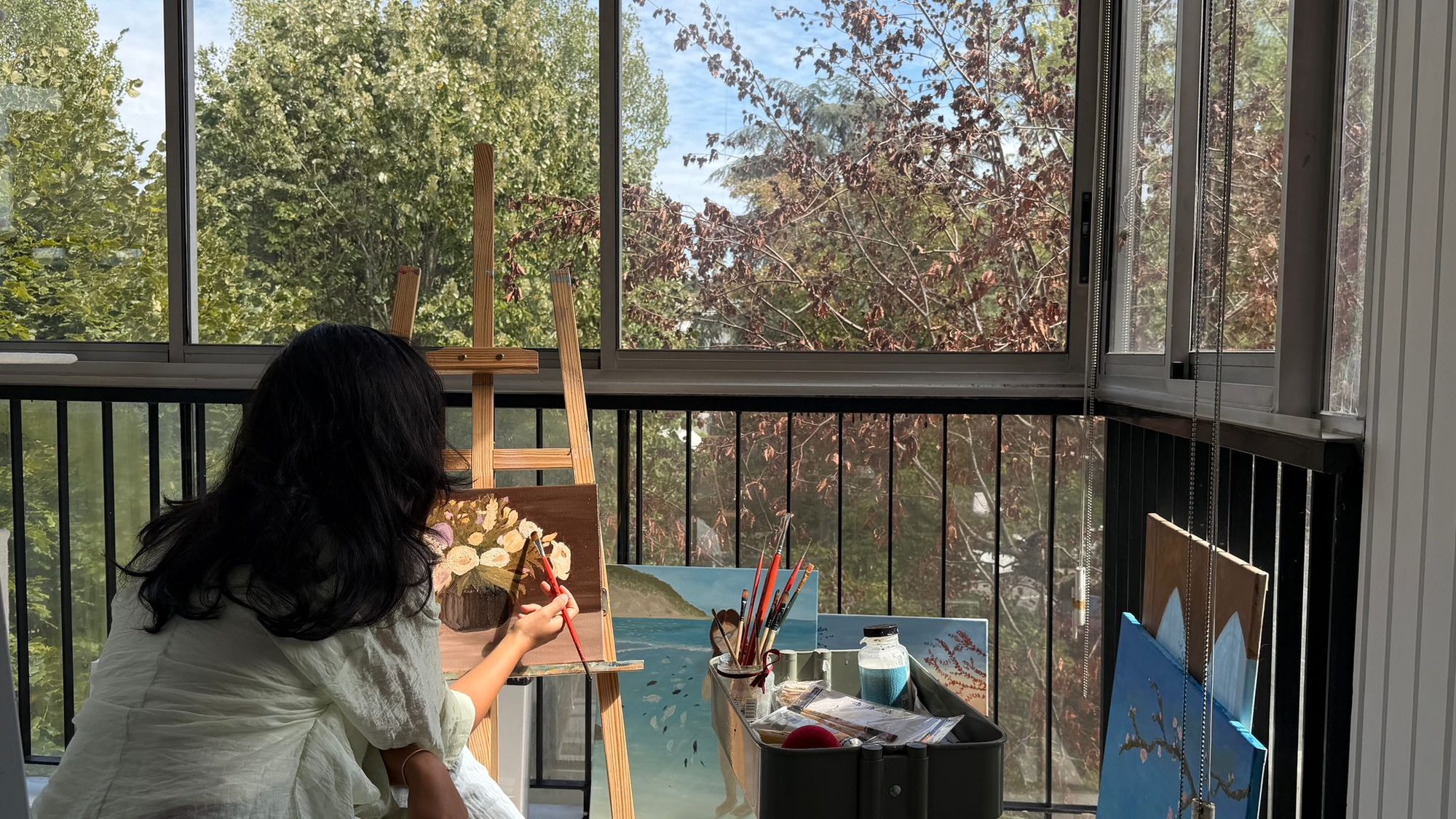Yous painting a still life at an easel on a sunlit balcony, with finished canvases resting against the railing